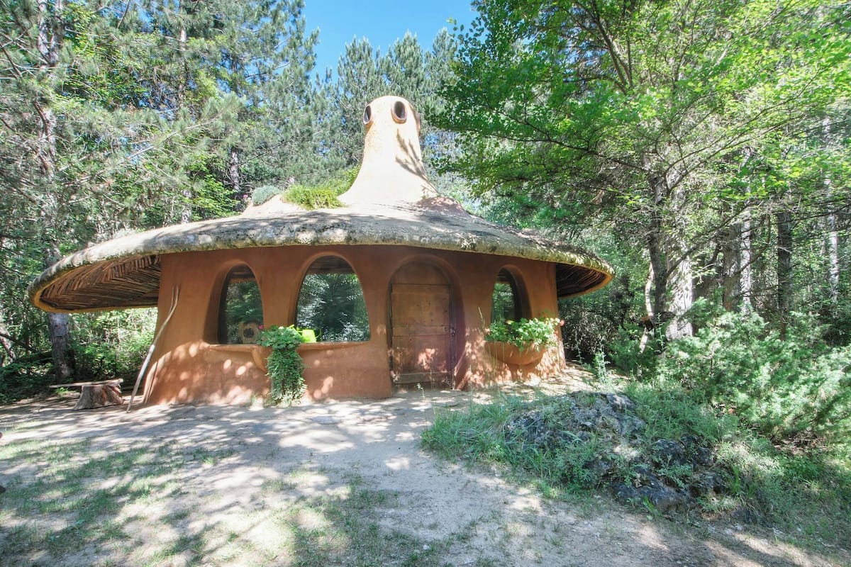 Un bâtiment en forme de champignon, doté d’une porte centrale et de fenêtres en arc de cercle, entouré d’une forêt.