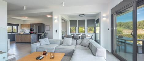 A modern open-plan living area with a sectional sofa, a wooden coffee table, and a dining table with chairs. There is a kitchen in the background, a balcony with a view, and large windows.