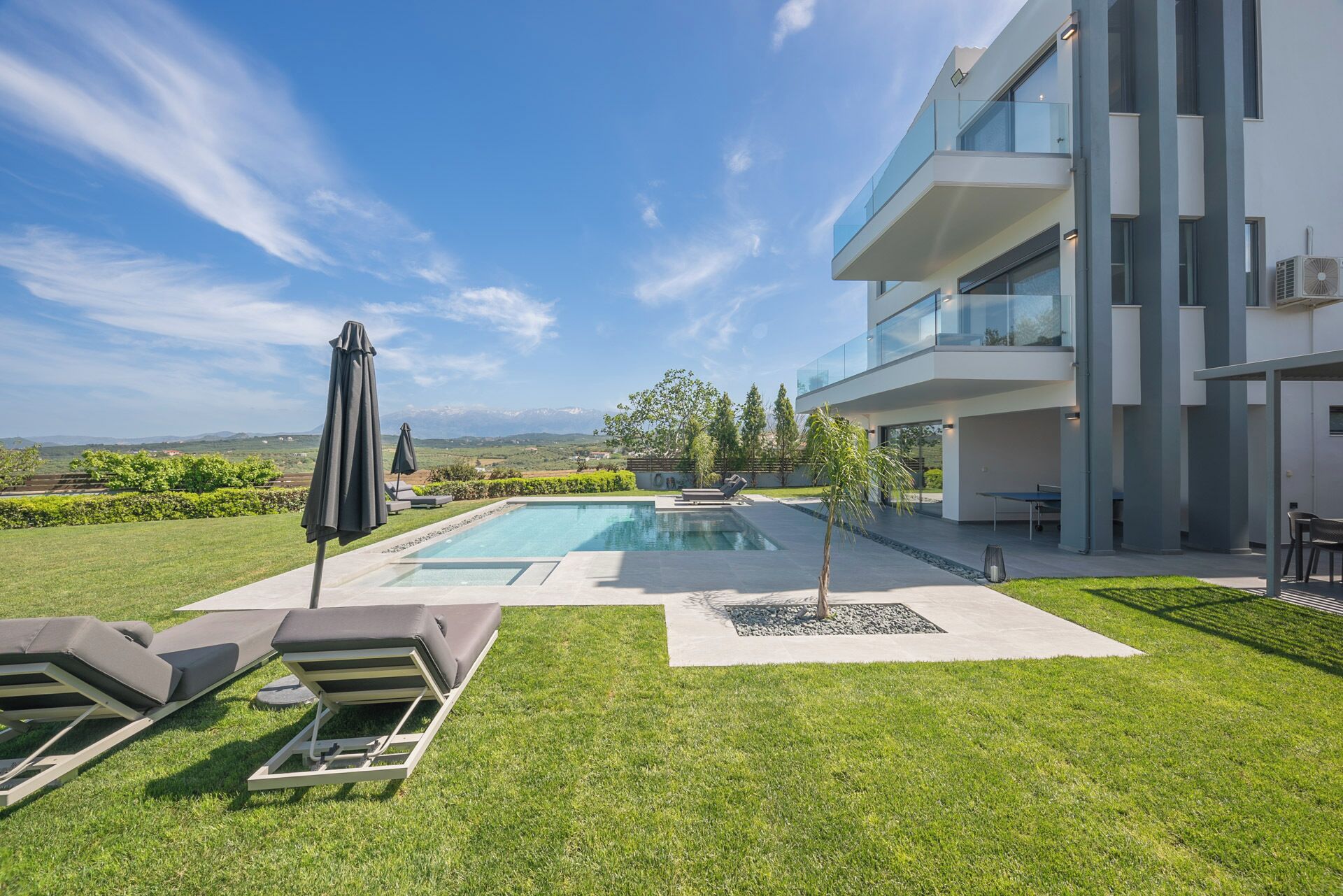 A modern outdoor pool area with lounge chairs, a covered umbrella, and a view of mountains in the background.