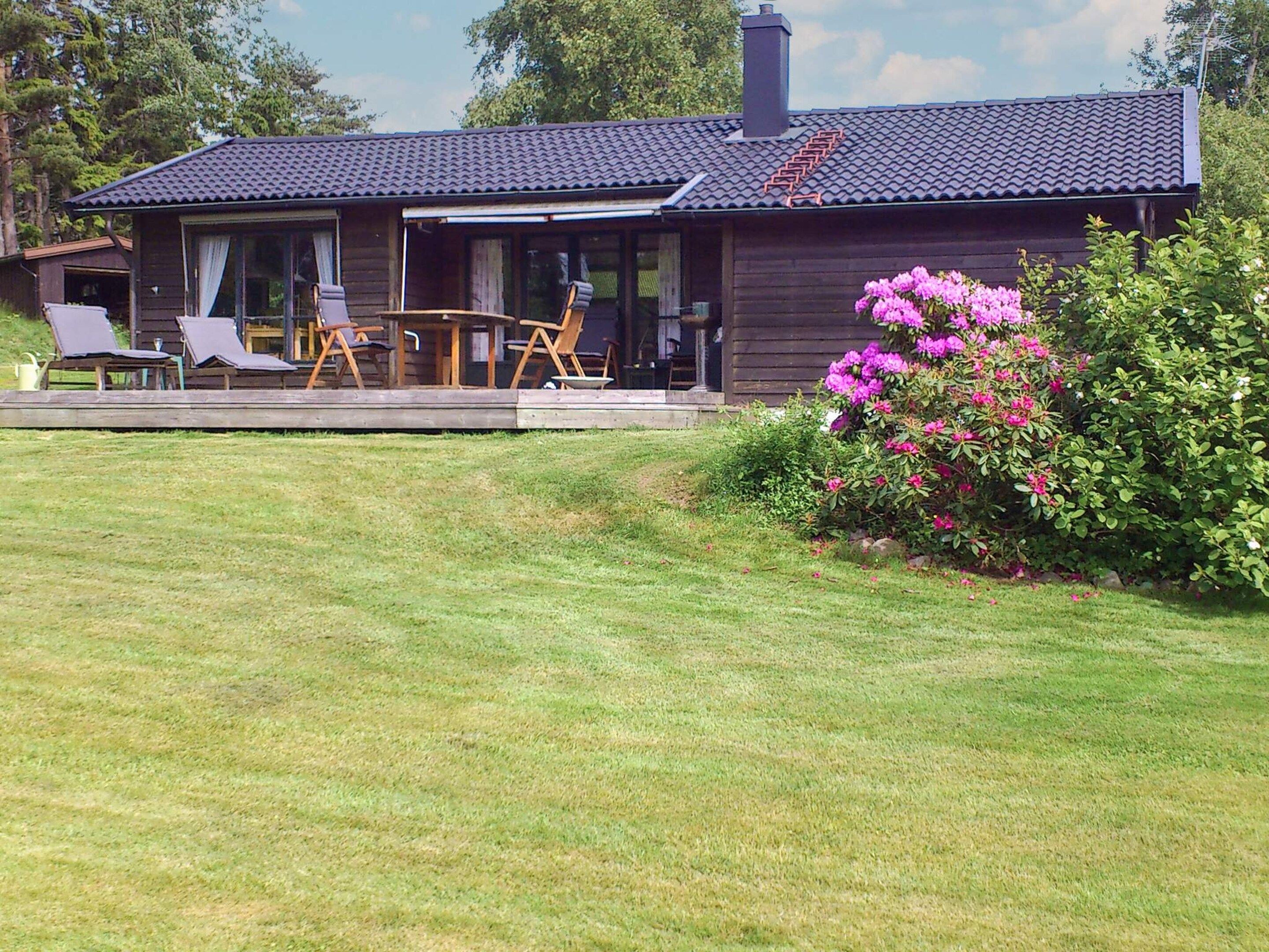 A wooden house with a tiled roof, a porch with chairs, and a garden with pink flowers.