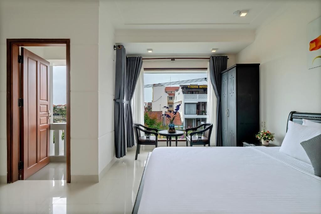 Habitación de hotel con cama, una pequeña mesa con sillas, un balcón con vistas a edificios y una puerta que da a otra habitación.