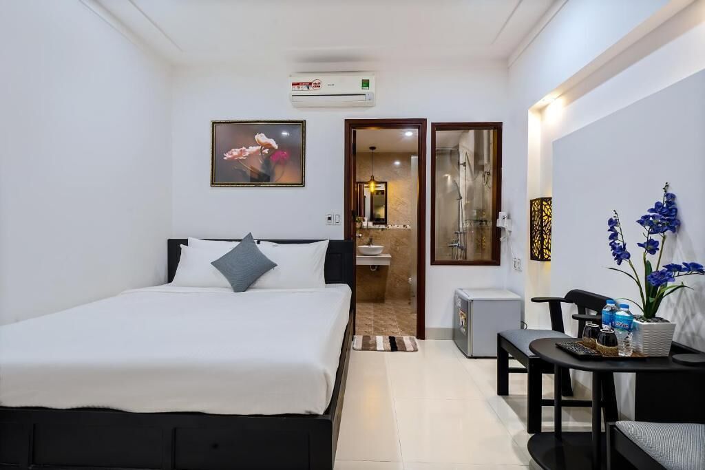 A hotel room with a bed, a desk, a chair, a small table with a vase of flowers, and a bathroom visible through an open door.
