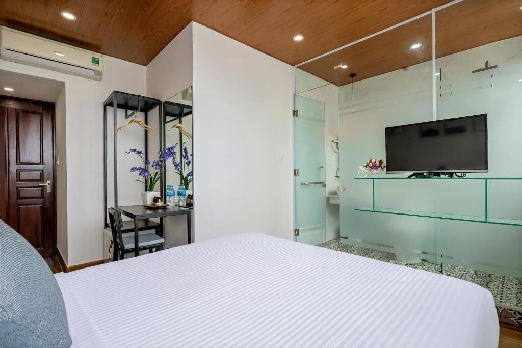 A hotel room with a bed, a desk with a chair, a TV on a glass cabinet, and a shower area.