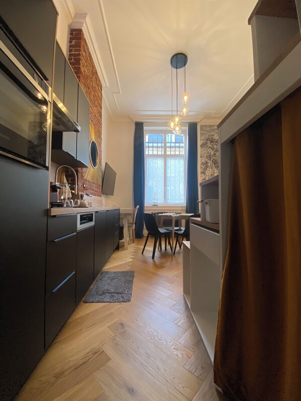 A modern kitchen with dark cabinetry, a dining area with a table and chairs, and a large window with curtains. - Studio in the Conciergerie of the former Banque de France in Flers. (Flers)