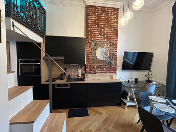 Una cocina moderna con pared de ladrillo decorativa, un área de comedor con mesa de vidrio y un televisor montado en la pared.