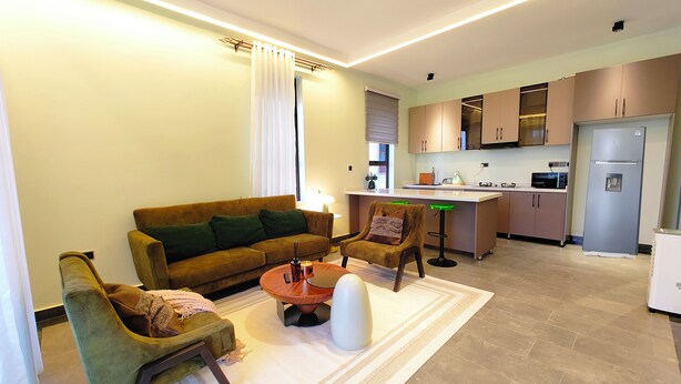 A modern living room with a sofa, chairs, a coffee table, and a kitchen in the background. - Heritage Residences-Urban Ranger 1BR (Kampala)