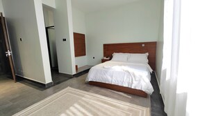 A hotel room with a large bed, a wooden headboard, a white bedspread, and a beige rug. - Heritage Residences-Urban Ranger 1BR (Kampala)