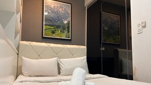 Una cama bien tendida con sábanas blancas y una toalla enrollada, un cuadro de un paisaje montañoso encima, y una puerta con un espejo en la pared contigua.