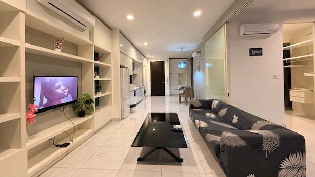 Una sala de estar moderna con un televisor de pantalla plana, una mesa de centro negra y un sofá con estampado de hojas. Hay estantes blancos con objetos decorativos y una unidad de aire acondicionado en la pared.