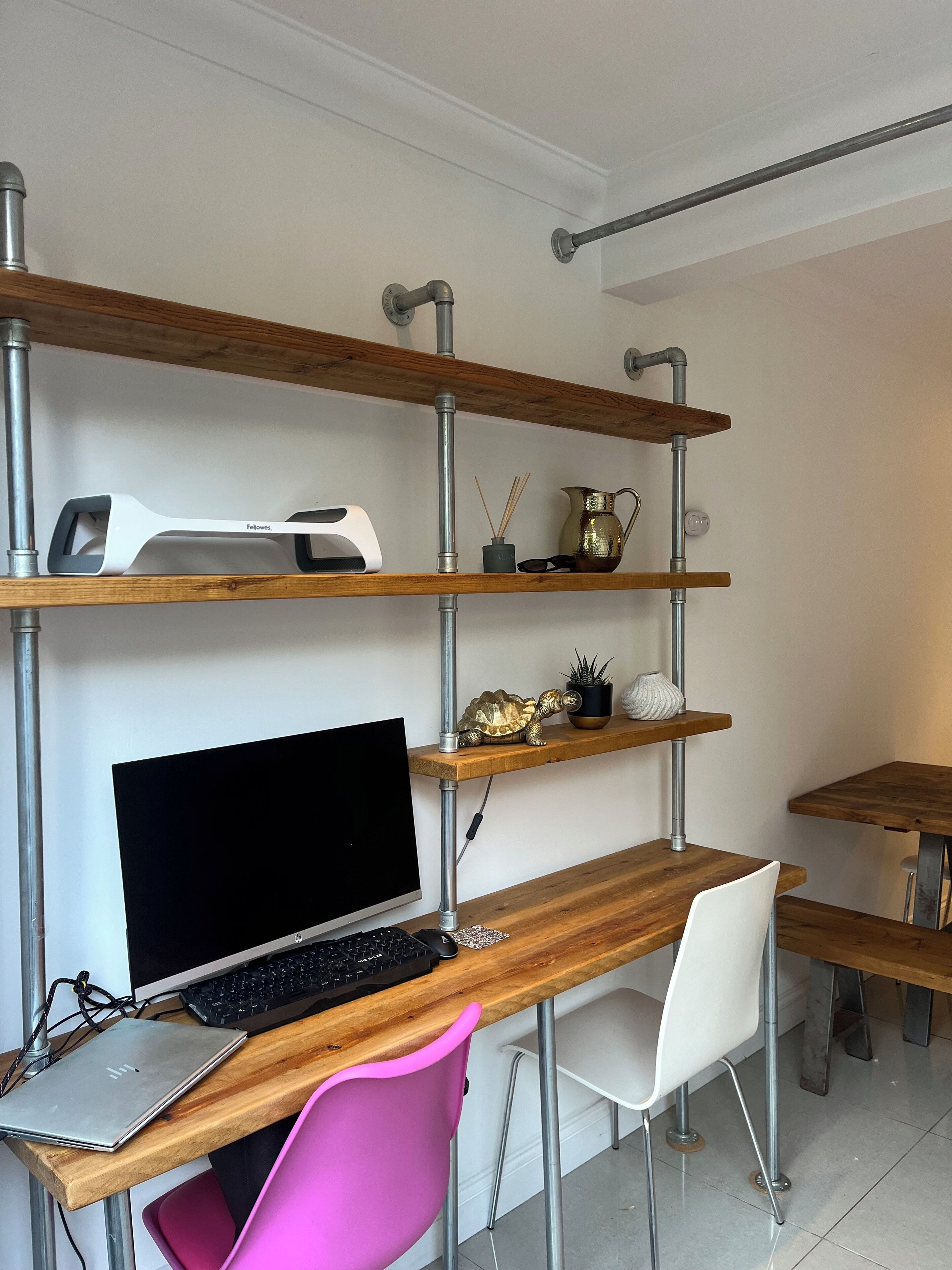 Un espacio de trabajo moderno con un escritorio de madera, una computadora y una silla rosa. Estantes superiores exhiben objetos decorativos y una lámpara.