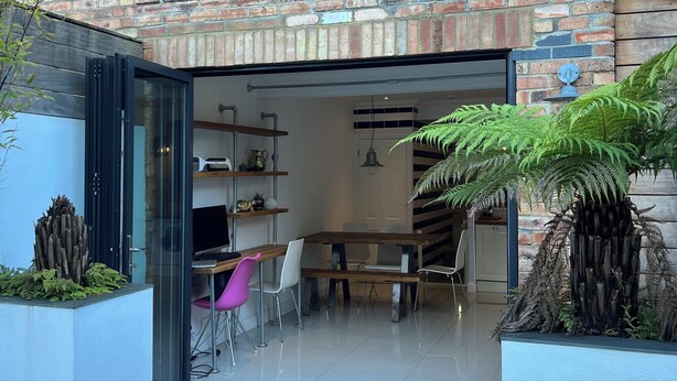 A modern brick building with a glass door entrance leading to an interior space with a dining table, chairs, and a fern plant.