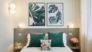 Un lit bien fait, agrémenté d’oreillers verts, une table de chevet avec une lampe et, au mur, des œuvres d’art encadrées représentant des feuilles tropicales.