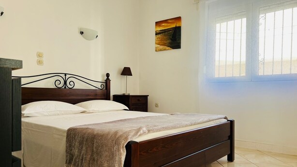 Una cama bien hecha con cabecera de madera, una mesita de noche con lámpara, un cuadro en la pared y una ventana con barrotes.