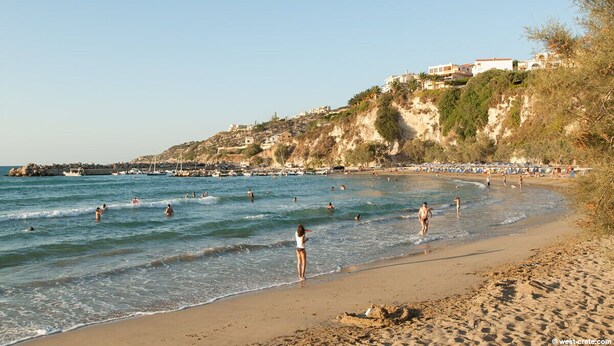 Uma praia com pessoas nadando e caminhando na orla, céu claro e edifícios ao fundo, numa encosta.