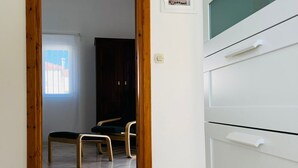 Una habitación con una puerta de madera que da a otra estancia, un armario blanco con cajones y un suelo de baldosas.