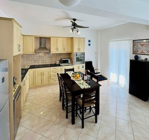A modern kitchen with light wood cabinets, a dining table with chairs, a ceiling fan, and a black cabinet. - LP Almyrida - 7 minutes walk from the beach (Almyrida)