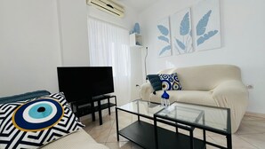Uma sala de estar com uma televisão de tela plana, uma mesa de centro de vidro, um sofá bege e uma almofada estampada.