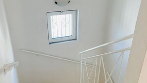 Uma escada com corrimão branco, uma pequena janela com grades decorativas e um luminário na parede.