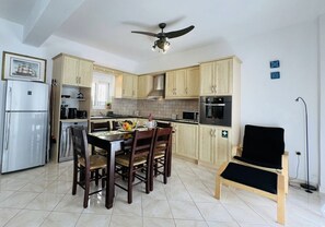 A compact kitchen with wooden cabinets, a dining table with chairs, a ceiling fan, and a black chair. - LP Almyrida - 7 minutes walk from the beach (Almyrida)