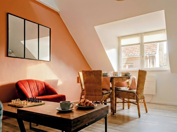 Ein gemütliches Wohnzimmer mit einem roten Sofa, einem Couchtisch aus Holz mit Schachfiguren, Korbstühlen und einem Esstisch mit Tellern und Gläsern.