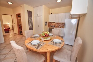 A dining area with a round wooden table set for four, surrounded by chairs. There is a kitchen area with white cabinets and a refrigerator to the right. - Daphne Two Bedroom Apartment (Corfu)
