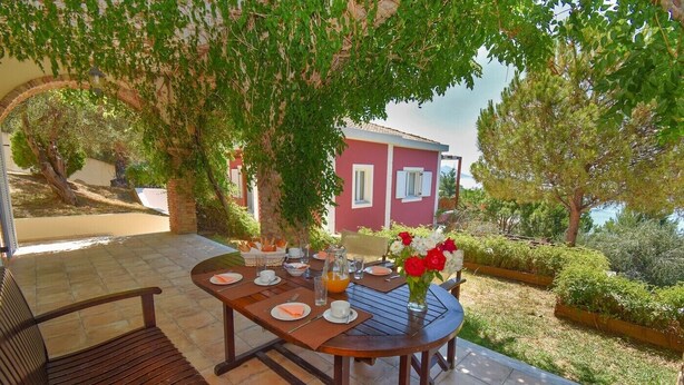 En terrasse med et rundt bord dækket til morgenmad, omgivet af grøn vegetation og en rød bygning i baggrunden.
