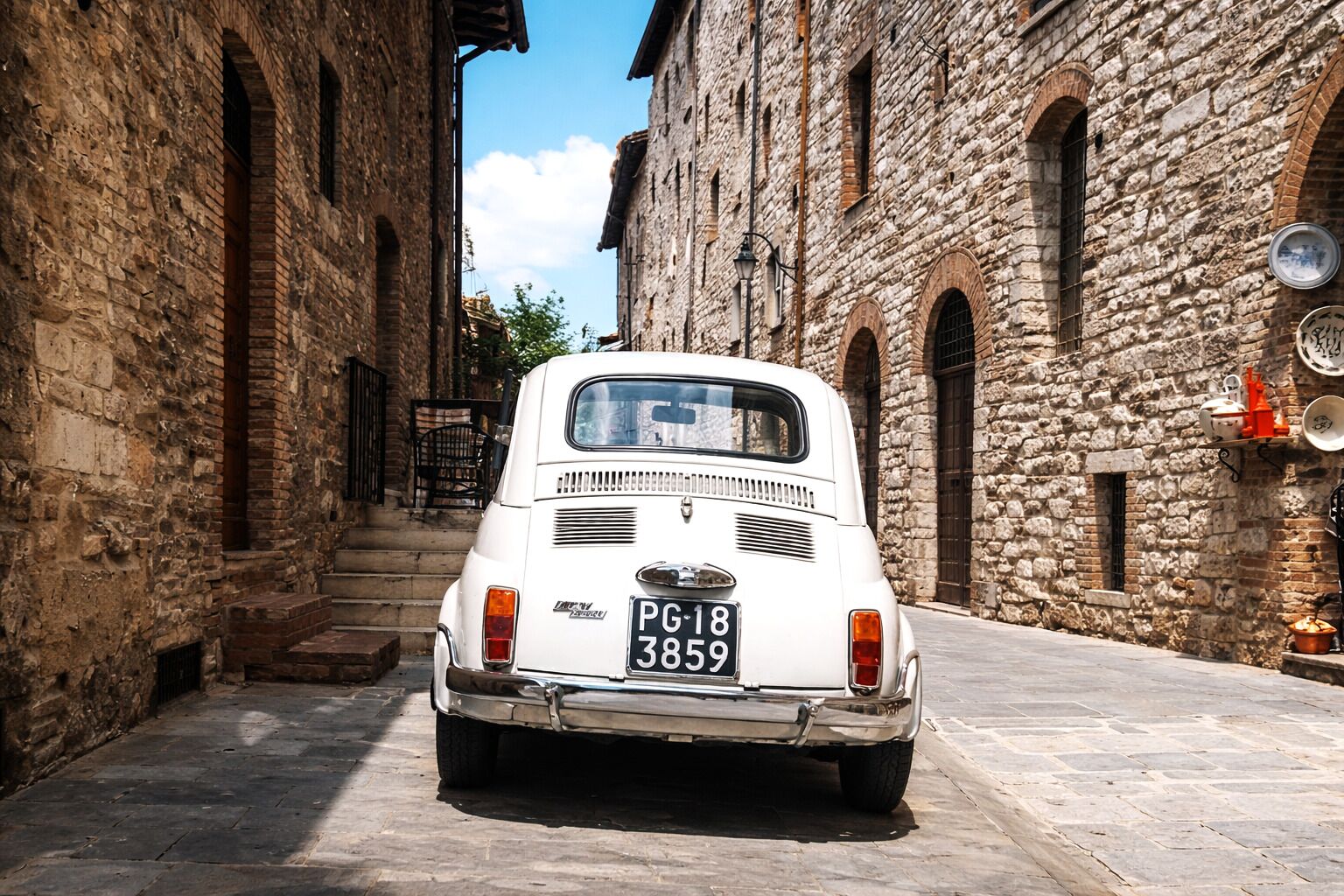 A vintage white car parked on a cobblestone street between stone buildings.