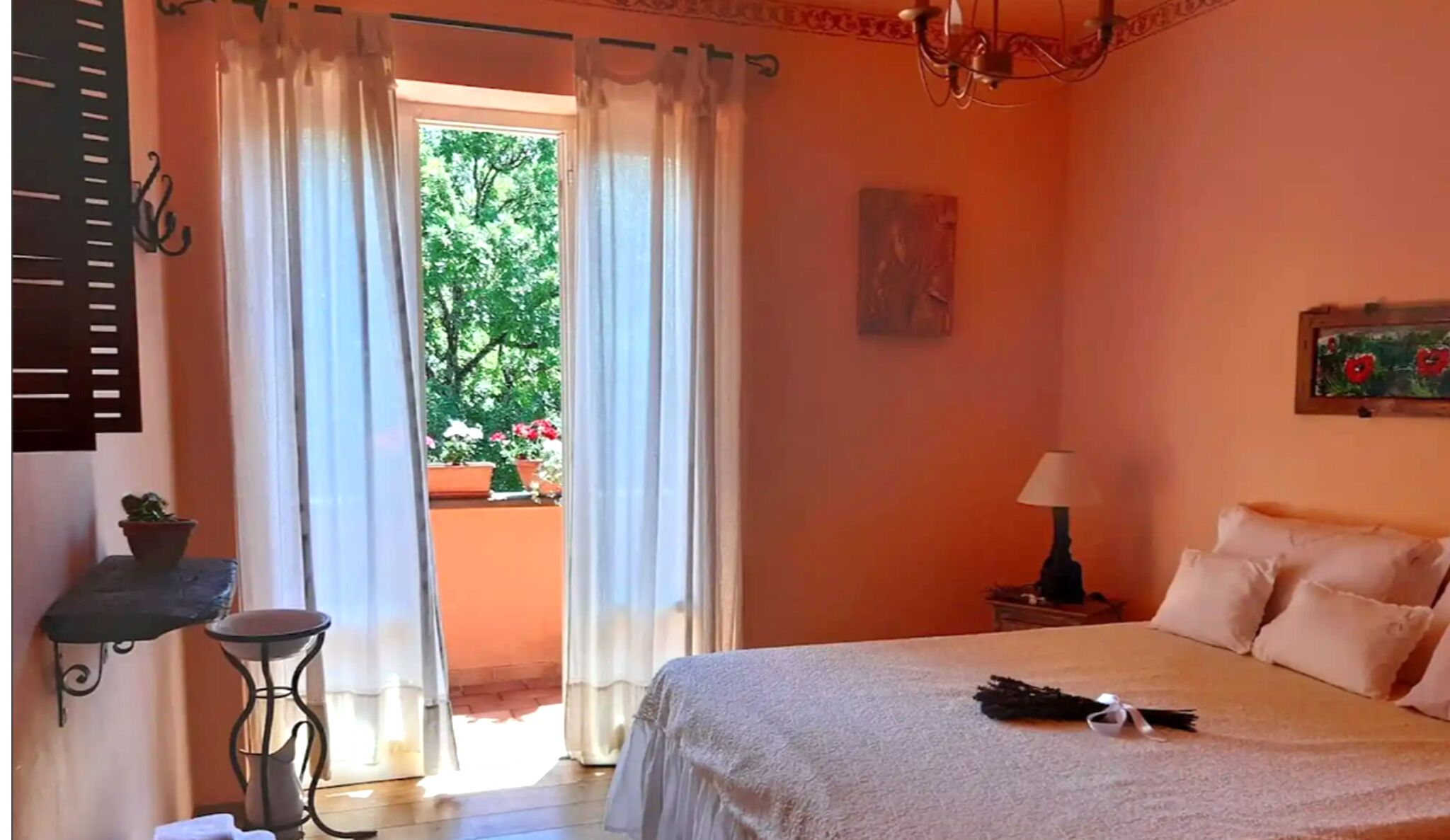 A bedroom with a bed, a nightstand, a lamp, a window with curtains, and a view of greenery outside.