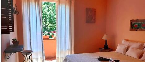 A bedroom with a bed, a nightstand, a lamp, a window with curtains, and a view of greenery outside.