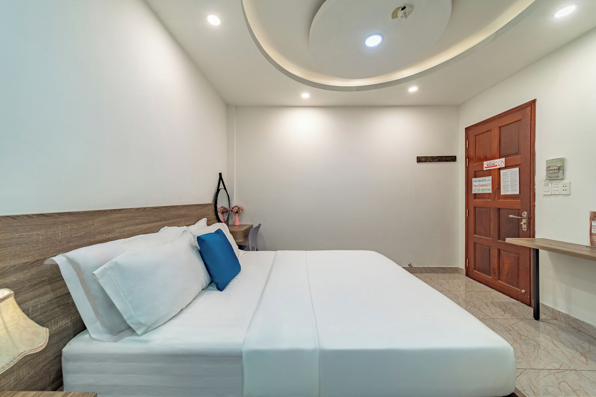 A neatly arranged hotel room with a bed, a nightstand, a wooden headboard, a door, and a wall-mounted shelf.