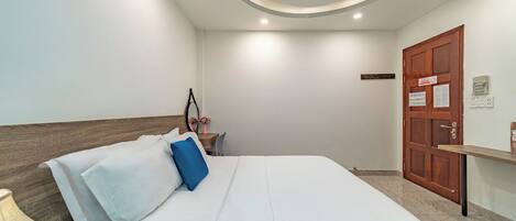 A neatly arranged hotel room with a bed, a nightstand, a wooden headboard, a door, and a wall-mounted shelf.
