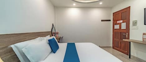 Una cama bien hecha con ropa de cama blanca y azul, un cabecero de madera, una mesita de noche con un florero y una puerta de madera con un letrero.