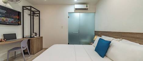Una camera d'albergo con un letto, una scrivania con un portatile, una sedia, una ventola da terra e un condizionatore d'aria.