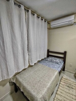 A bedroom with a bed, curtains, an air conditioning unit, and a patterned rug. - Ground floor one-bedroom apartment in a condominium with swimming pools and 100 meters from the sea in Florianópolis, SC. (Florianópolis)
