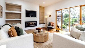 A modern living room with a fireplace, a television, and a large sliding glass door leading to a garden. - The Greenhouse - Spa & Fire - Pet Friendly (Barwon Heads)