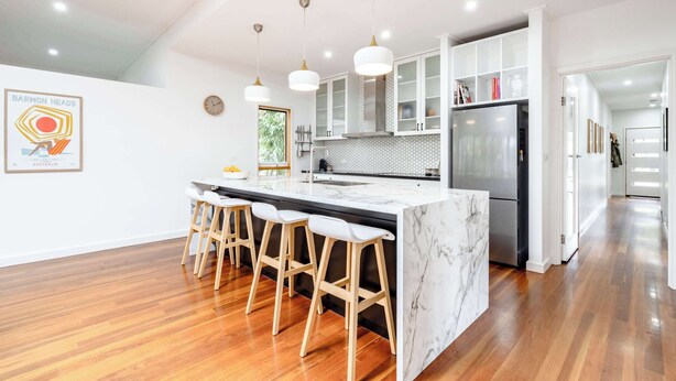 A modern kitchen with a marble island, wooden stools, pendant lights, and a stainless steel refrigerator. - The Greenhouse - Spa & Fire - Pet Friendly (Barwon Heads)