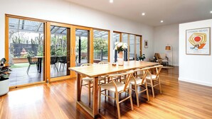 A spacious dining area with a wooden table and chairs, large sliding glass doors leading to an outdoor patio, and a framed artwork on the wall. - The Greenhouse - Spa & Fire - Pet Friendly (Barwon Heads)