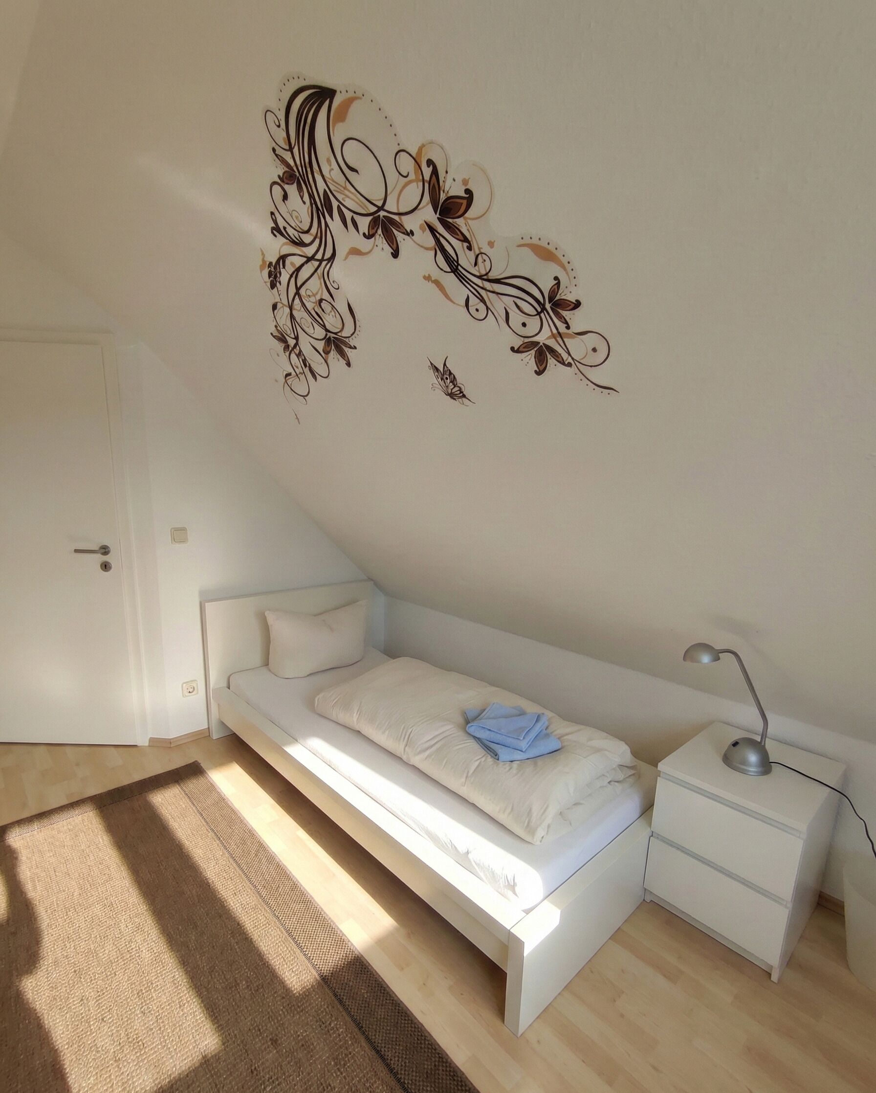 A modern bedroom with a white bed, a bedside table with a lamp, and a decorative wall piece.