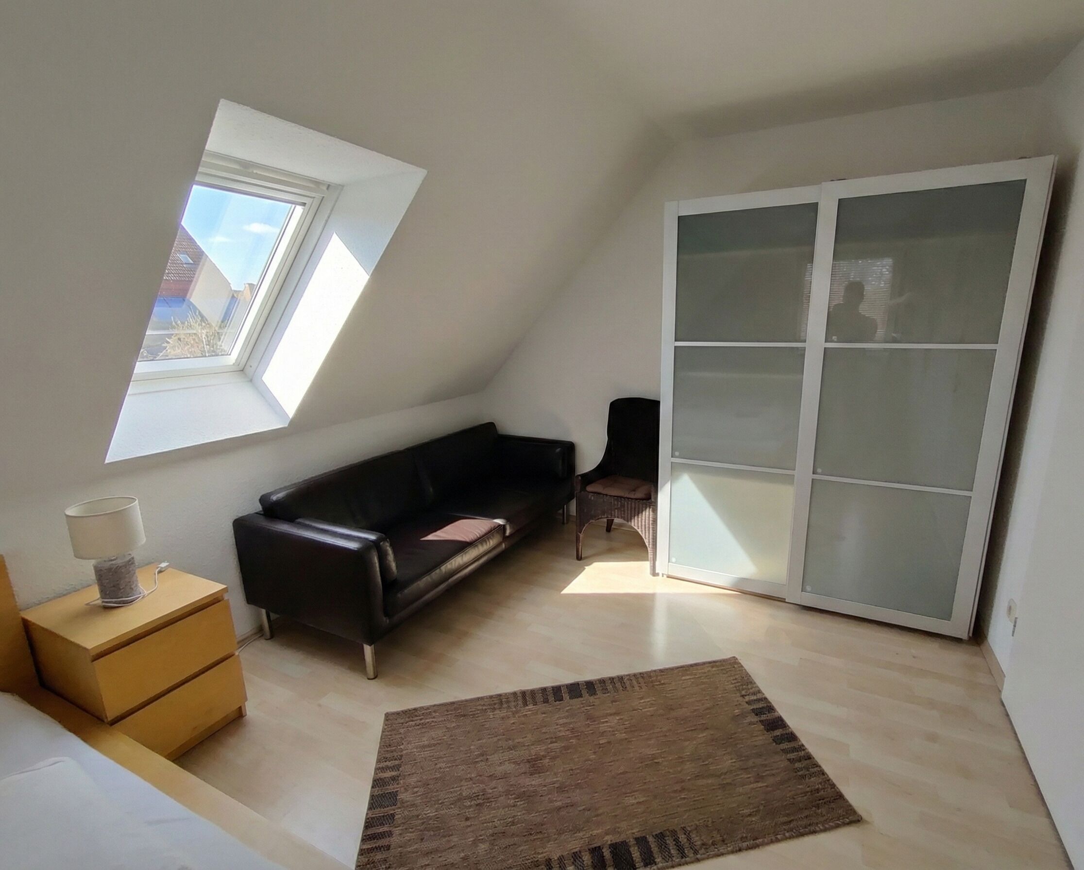 Una habitación con techo inclinado, un sofá de cuero negro, una pequeña mesa de madera con una lámpara, una alfombra marrón y un ventanal corredero.