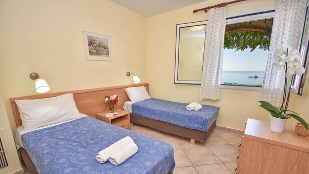 Ein Hotelzimmer mit zwei Betten, einem Holzkopfteil, einem Nachttisch mit Lampe, einem kleinen Tisch mit Blumenvase, einem Fenster mit Meerblick und einem Schreibtisch mit Pflanze.