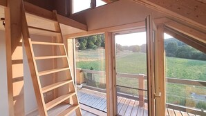 Une chambre à coucher aménagée dans un grenier, avec une échelle en bois, une grande porte-fenêtre coulissante et une vue sur un paysage verdoyant.