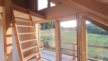 A loft bedroom with a wooden ladder, a large sliding glass door, and a view of a green landscape.