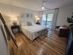 Ein Schlafzimmer mit Bett, Nachttischen, Deckenventilator, Fenster mit Vorhängen und Blick ins Freie.
