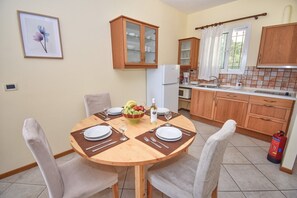 A dining area with a round wooden table set for two, surrounded by chairs. A fruit bowl and a bottle of wine are on the table. The room has wooden cabinets and a tiled backsplash in the kitchen area. - Aithra One Bedroom Apartment (Pelekas)