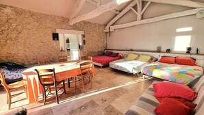 A cozy room with a stone wall, a wooden dining table with chairs, a bed with colorful bedding, and a sofa with red cushions.