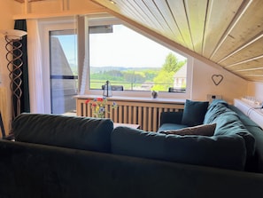Una habitación acogedora con un sofá verde, una ventana con vistas a la vegetación, una decoración con forma de corazón en la pared y una pequeña mesa con una linterna.