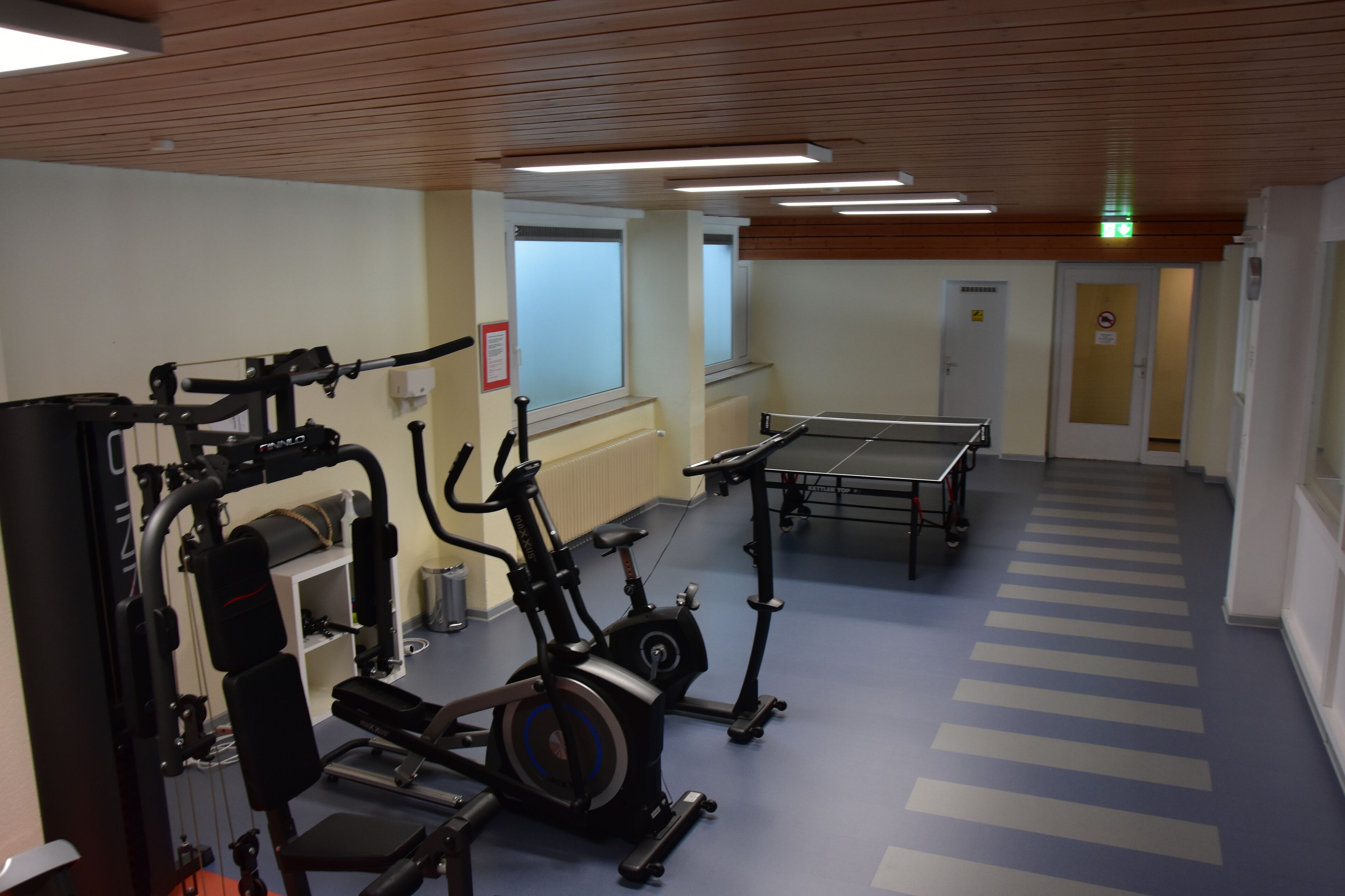 A gym with exercise equipment, a ping pong table, and a window with blinds.
