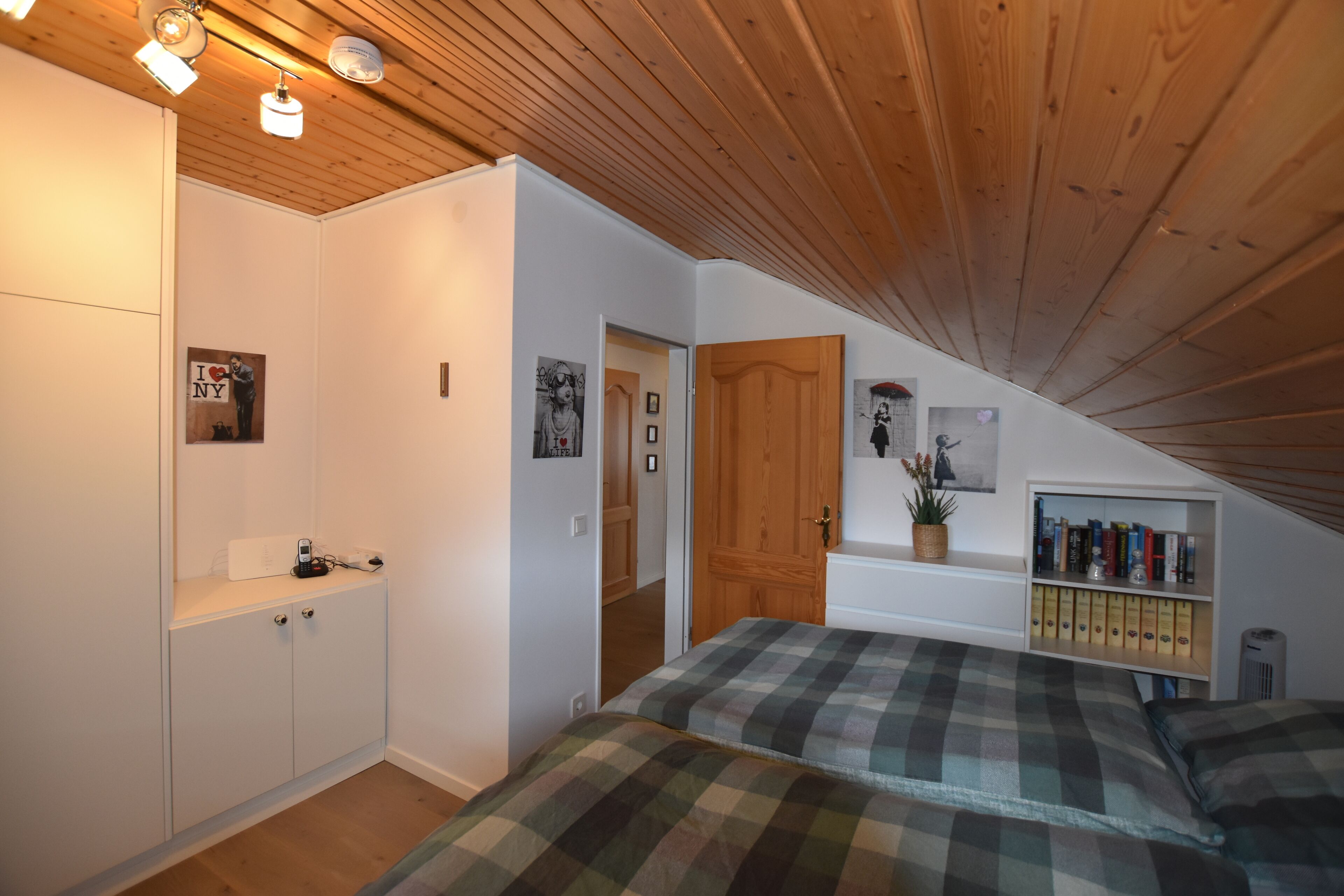 A bedroom with a sloped ceiling, a bed with plaid bedding, a bookshelf, and a door leading to another room.