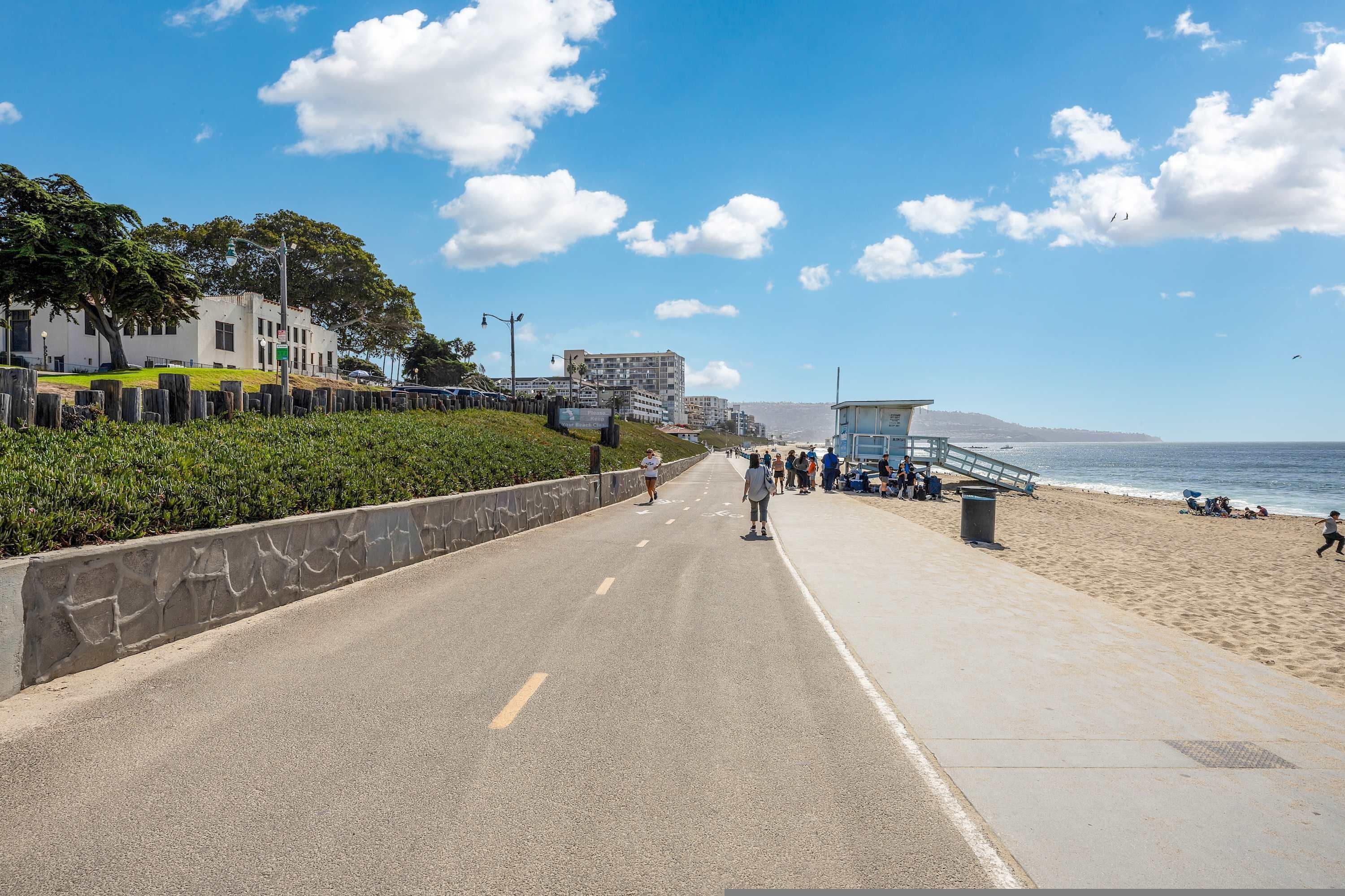 A coastal road with a beach on the right, a lifeguard tower, and a building with a green sign.