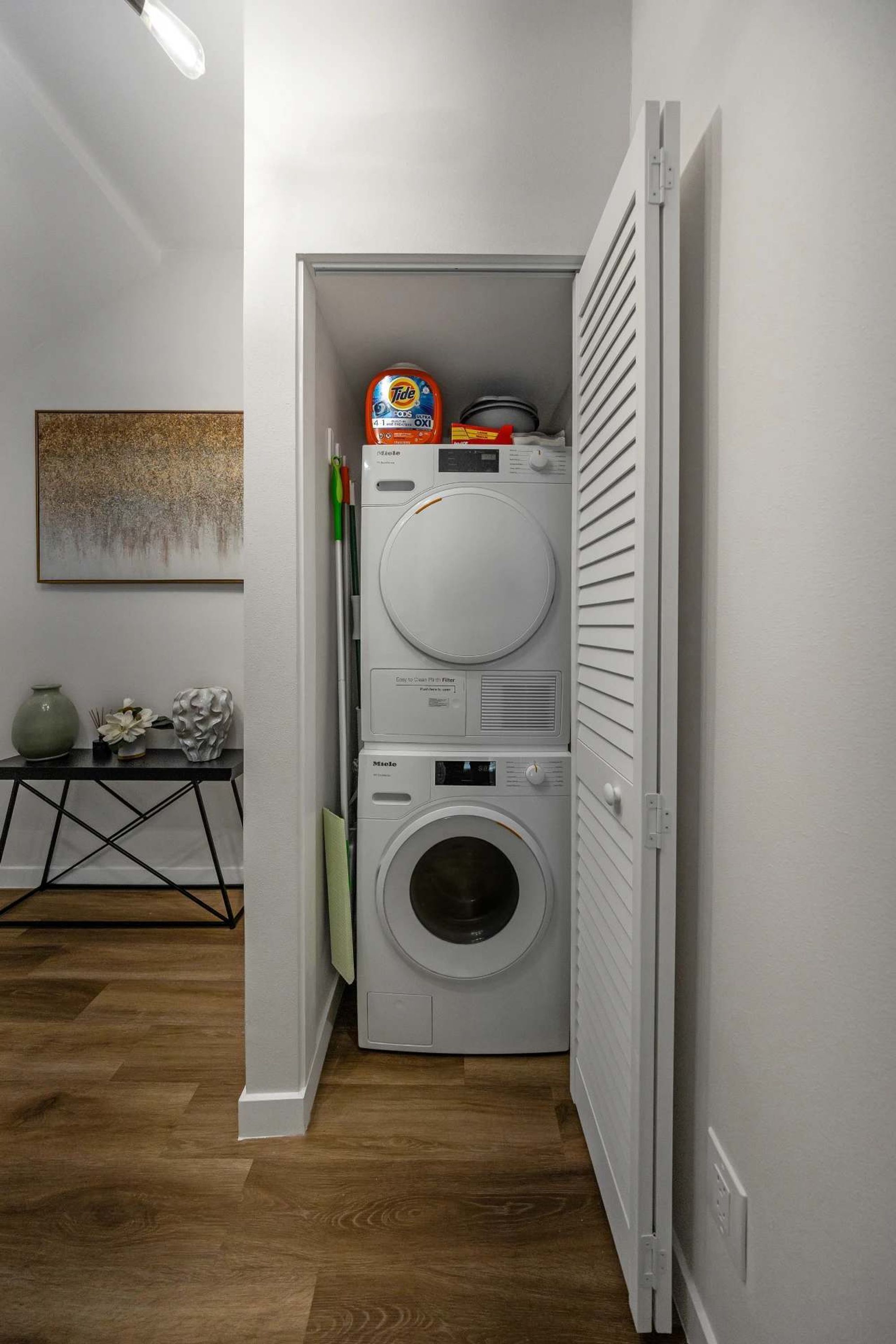 Lavandería con lavadoras y secadoras apiladas, una mesita con objetos decorativos y un cuadro en la pared.